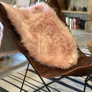 May include: A pink faux fur throw on a brown leather butterfly chair with a black metal frame. The chair is on a striped rug.