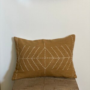 May include: A mustard yellow throw pillow with a white diamond pattern stitched on the front. The pillow is sitting on a wooden stool.