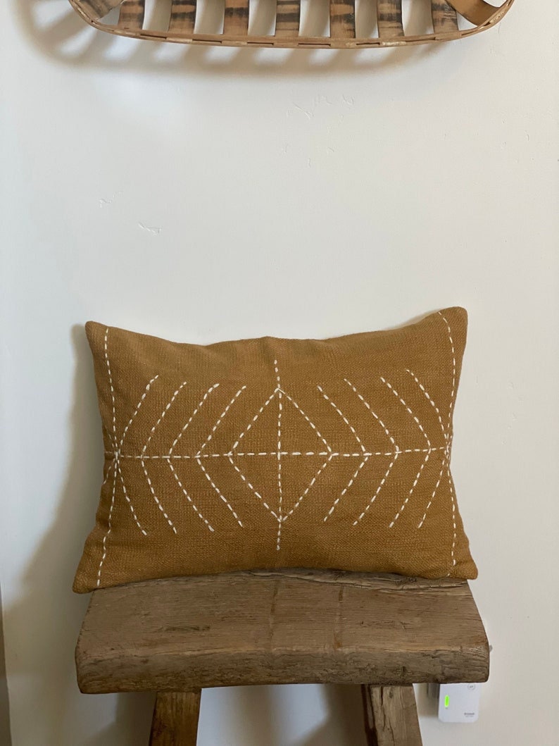 May include: A mustard yellow throw pillow with a white diamond pattern stitched on the front. The pillow is sitting on a wooden stool.