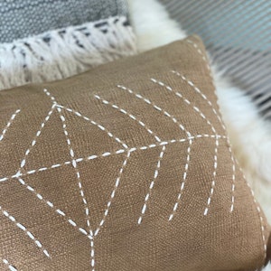 May include: A brown throw pillow with a white geometric design stitched on the front. The pillow is made of a textured fabric.