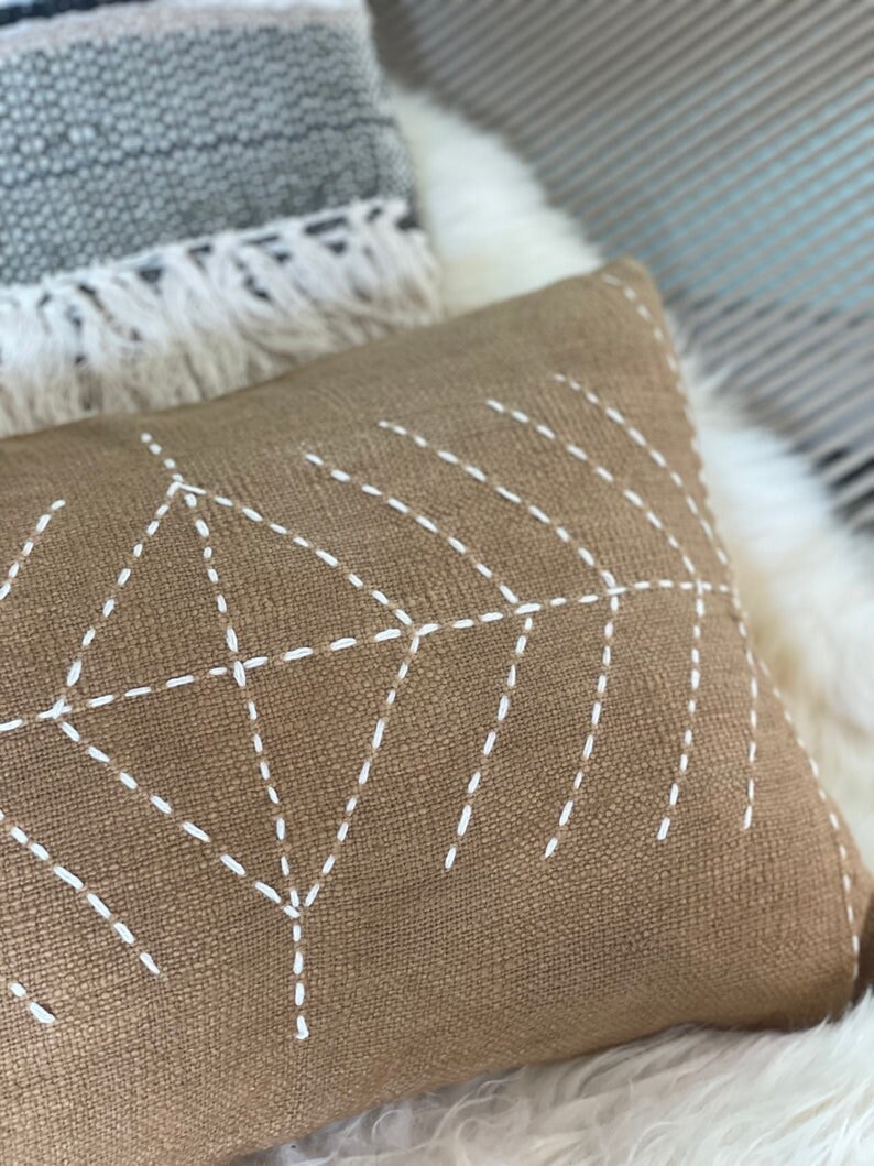 May include: A brown throw pillow with a white geometric design stitched on the front. The pillow is made of a textured fabric.