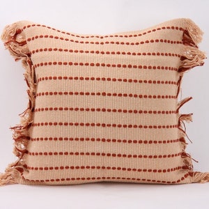 May include: A square throw pillow with a cream colored fabric and red stripes. The pillow has a fringe border.