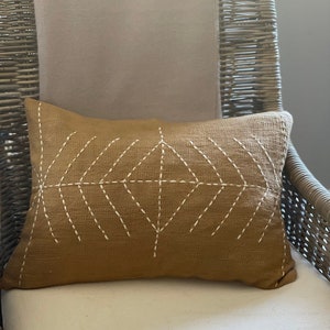 May include: A brown throw pillow with a white geometric design stitched on the front. The pillow is sitting on a wicker chair with a beige cushion.