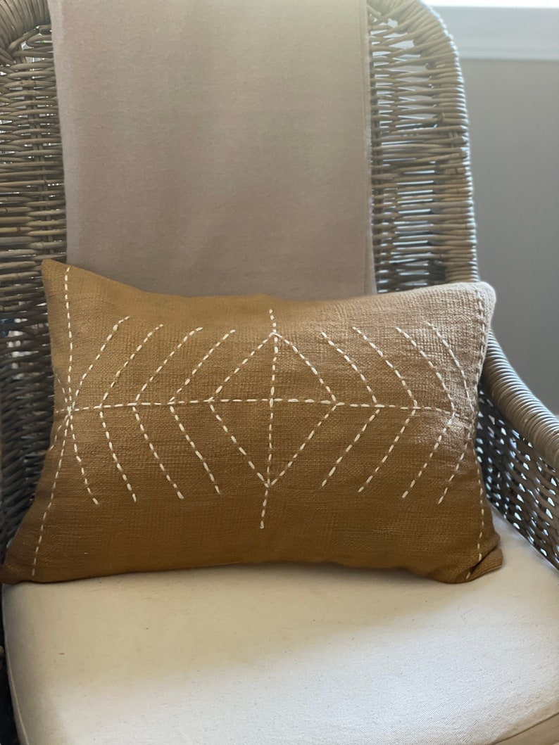 May include: A brown throw pillow with a white geometric design stitched on the front. The pillow is sitting on a wicker chair with a beige cushion.