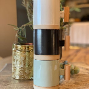 May include: Three ceramic mugs stacked on top of each other. The top mug is white, the middle mug is black, and the bottom mug is light green. Each mug has a wooden handle.