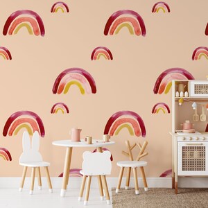 May include: A children's playroom with a peach-colored wall adorned with watercolor rainbow decals. The room features a white play kitchen, a small table, and chairs with animal-shaped backs. A woven basket and rug complete the scene.