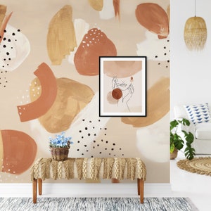May include: A beige wall with a brown, white, and black abstract pattern. A black framed print of a line drawing of a woman's face with hands on her cheeks is hanging on the wall. A small brown wicker basket with blue flowers sits on a light brown bench with fringe.