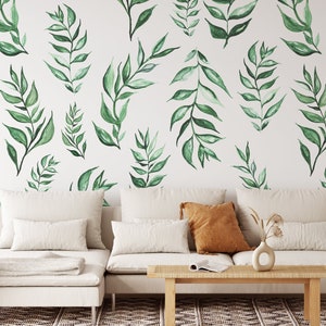 May include: A white couch with white throw pillows and a brown throw pillow sits in front of a white wall with a green watercolor leaf pattern. A small wooden side table with a woven rattan top is to the right of the couch. A small potted plant with dried beige stems sits on the floor in front of the couch.