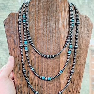 May include: Three-strand necklace set displayed on a wooden stand. The necklaces feature silver-colored beads and turquoise accents. The beads vary in size, with larger ones interspersed throughout the strands. The stand is a warm brown color.