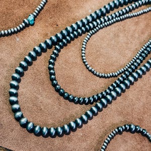 May include: Three strands of black and silver beaded necklaces on a brown suede surface. The necklaces are made of round beads with a smooth, polished finish.