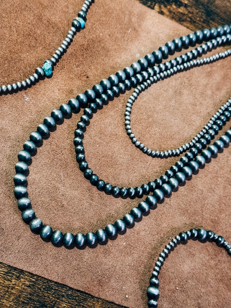 May include: Three strands of black and silver beaded necklaces on a brown suede surface. The necklaces are made of round beads with a smooth, polished finish.