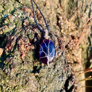 Op de afbeelding: Een handgemaakte hangerketting met een dieppaarse, ovale steen en een draadomwikkeld boomontwerp. De hanger hangt aan een zwart koord. De ketting staat tegen een getextureerde, bruine achtergrond. Een uniek sieraad.