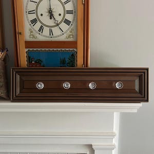 May include: A dark brown wooden wall-mounted coat rack with four clear crystal knobs. The rack is rectangular with a recessed panel design. A wooden clock with a white face and black Roman numerals is in the background.
