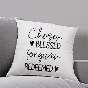 Könnte beinhalten: Ein weißes Kissen mit schwarzem Text, der "Chosen Blessed Forgiven Redeemed" lautet, mit einem Herzsymbol vor "Blessed" und nach "Redeemed".