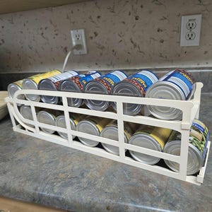 May include: White metal can rack with multiple rows of canned goods. The cans are labeled with various food items, including beans and vegetables.