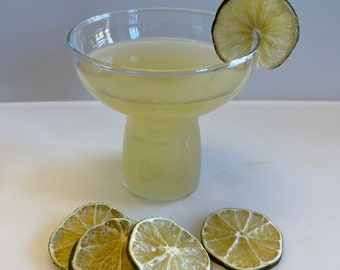 Pode incluir: Um copo de cocktail transparente cheio de líquido amarelo, decorado com uma fatia de limão. Quatro fatias de limão secas estão dispostas sobre uma superfície branca ao lado do copo. O copo tem uma taça larga e uma base curta.