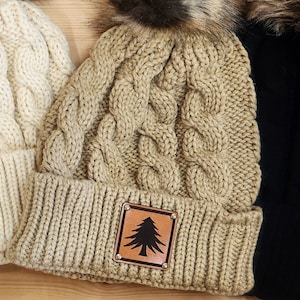 May include: A tan cable-knit beanie with a faux fur pom-pom. The hat has a brown leather patch with a black tree design. The ribbed brim and the body of the hat are in a matching tan colour. The hat is displayed with other beanies.