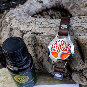 May include: A silver aromatherapy diffuser featuring a tree of life design and a brown leather strap. An essential oil bottle with a green label and black cap is next to the diffuser. The diffuser has an orange interior.