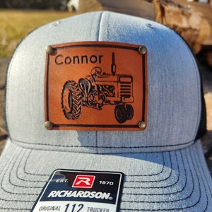 Op de afbeelding: Grijze truckerpet met een zwarte mesh achterkant en een bruine leren patch. De patch toont de naam "Connor" boven een gegraveerd tractorontwerp. De pet bevat een "Richardson" label.