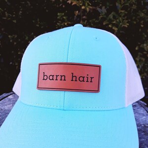 Puede incluir: Gorra de camionero turquesa con la parte trasera de malla blanca. Un parche de cuero marrón en la parte delantera muestra las palabras "barn hair". El sombrero está sobre una superficie de madera.