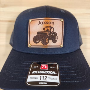 Op de afbeelding: Marineblauwe truckerpet met een leren patch met een tractorafbeelding en de naam "Jaxson". De pet heeft een mesh achterkant en een gebogen klep. Het Richardson-label luidt "Original 112 Trucker Youth".
