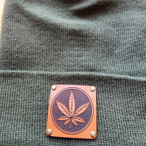 Op de afbeelding: Een olijfgroene gebreide beanie met een vierkante, bruine leren patch. De patch heeft een cirkelvormig ontwerp met een gedetailleerd cannabisblad. De patch is bevestigd met vier kleine, messingkleurige klinknagels.