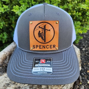 Può includere: Cappellino da camionista grigio e bianco con una toppa in pelle raffigurante un elettricista e il nome "Spencer". Il cappellino ha una visiera curva e un cinturino regolabile. L'etichetta del marchio Richardson è visibile.