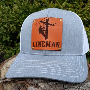 May include: A grey trucker cap with a white mesh back and a curved peak. A brown leather patch on the front displays a lineman silhouette on a utility pole, with the word "LINEMAN" beneath. The patch is secured with brass rivets.