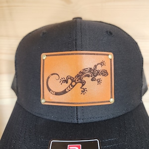 May include: Black trucker hat with a brown leather patch featuring a detailed lizard design. The hat has a mesh back and a curved brim. The Richardson brand tag is visible at the bottom.