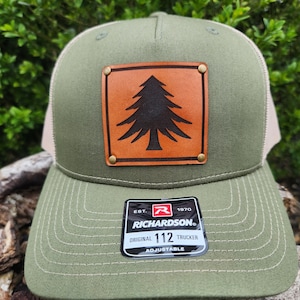 May include: Olive green trucker cap with a tan mesh back and a brown leather patch featuring a black tree design. The cap has a curved peak and a Richardson 112 Trucker tag.