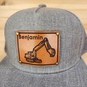 May include: Gray trucker hat with a brown leather patch. The patch features the name "Benjamin" above an excavator illustration. The patch is secured with brass rivets. The hat has a mesh back.