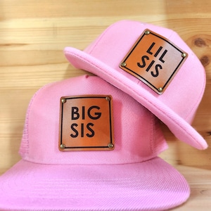Puede incluir: Dos gorras de béisbol rosas con parches de cuero marrón. Un parche dice "BIG SIS" y el otro "LIL SIS" en letras negras. Las gorras tienen una visera plana y una parte trasera de malla.