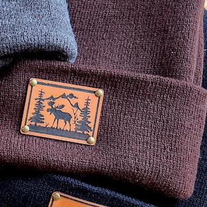 以下が含まれることがあります： グレー、バーガンディ、ネイビーブルーの3色のニット帽のクローズアップ。バーガンディのビーニーには、ヘラジカ、木、山々の黒いシルエットが描かれた長方形の革パッチが付いています。