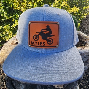 Puede incluir: Gorra de béisbol azul claro con visera plana. Cuenta con un parche de cuero marrón con una silueta negra de un piloto de motocross y una motocicleta. El nombre "MYLES" está impreso debajo.