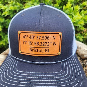 May include: Navy blue trucker hat with a white mesh back and a curved brim. A brown leather patch is affixed to the front, displaying the coordinates 41° 40' 37.596" N, 71° 15' 58.3272" W, and the text "Bristol, RI".