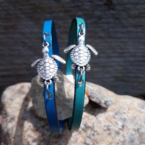 Könnte beinhalten: Zwei Lederarmbänder, eines blau und eines türkis, jeweils mit einem silberfarbenen Schildkrötenanhänger. Die Armbänder werden auf einem Felsen mit einem hölzernen Hintergrund präsentiert.