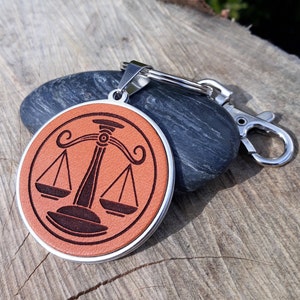 Op de afbeelding: Een ronde, bruin lederen sleutelhanger met een zilveren rand en sluiting. De sleutelhanger heeft een zwarte afbeelding van de weegschaal van de rechtvaardigheid. De achtergrond bevat een donkergrijze steen en een houten oppervlak.