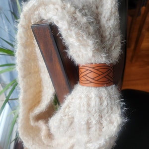May include: Cream-coloured infinity scarf made from thick, textured yarn. A brown leather cuff with an etched geometric design holds the scarf in place. The scarf is draped over a wooden chair.