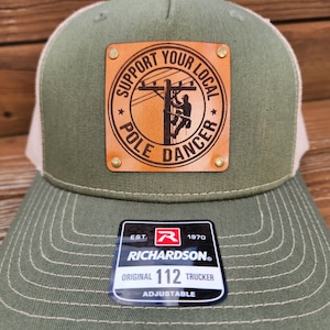 May include: An olive green trucker hat with a tan mesh back. The front has a brown leather patch with the words "SUPPORT YOUR LOCAL POLE DANCER" around a lineman image. The hat includes a Richardson 112 tag.