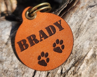 May include: A round, orange leather pet tag with the name "BRADY" embossed in dark brown letters. Two paw prints are also embossed on the tag. A brass ring is attached at the top for easy attachment to a collar.