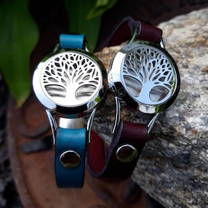 May include: Two aromatherapy bracelets with stainless steel tree of life pendants. One has a teal leather strap, the other a burgundy leather strap. The bracelets are displayed on a rock surface.