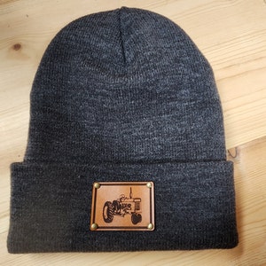 Op de afbeelding: Donkergrijze gebreide beanie met een bruin leren patch met een tractorillustratie. De patch is bevestigd met vier messingkleurige klinknagels. De hoed is gemaakt van zacht gebreid materiaal.