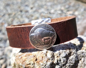 May include: Dark brown bison hide leather cuff bracelet with a silver buffalo nickel. The bracelet has a simple design with a metal clasp. The text "Dark Brown - Bison Hide" is visible.