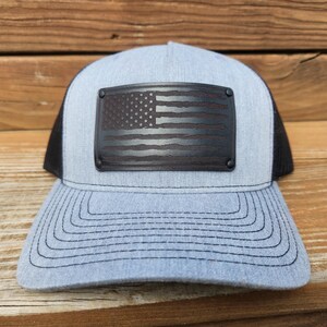 May include: A light blue trucker hat with a black mesh back and a curved brim. The front features a dark gray rectangular patch with a distressed American flag design. The hat has a snapback closure for an adjustable fit.