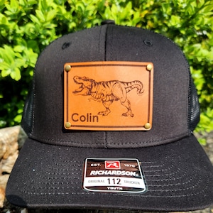 Puede incluir: Gorra de camionero negra con un parche de cuero marrón con una ilustración de Tyrannosaurus Rex y el nombre "Colin". La gorra tiene una parte trasera de malla y una visera curva. La etiqueta Richardson es visible.