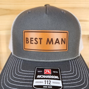 May include: Gray trucker hat with a white mesh back and a brown leather patch that reads "BEST MAN". The hat is a Richardson 112 Trucker, one size fits all.