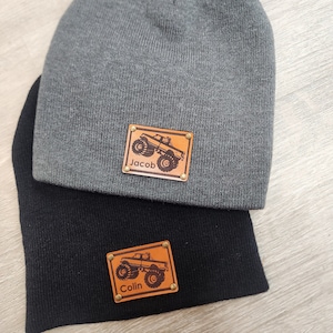 May include: Two beanies, one charcoal grey and one black, are stacked. Each beanie has a rectangular leather patch with a monster truck graphic and a name. The grey beanie's patch says "Jacob", and the black beanie's patch says "Colin".