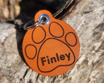 May include: A round, orange leather pet tag with a paw print design and the name "Finley" engraved in black. The tag has a silver metal ring for attachment.