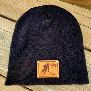 May include: A navy blue knitted beanie hat with a rectangular leather patch. The patch displays a hockey player illustration, the name "Colin," and the number "#17." The hat is made of a soft, knitted material.
