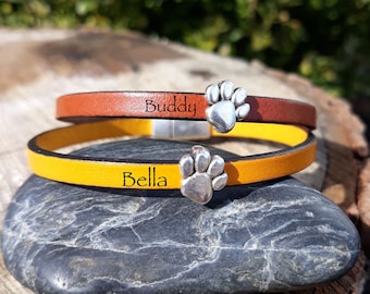 May include: Two leather bracelets, one brown and one yellow, are stacked on a rock. Each bracelet has a silver paw print charm. The brown bracelet is inscribed with the name "Buddy", and the yellow bracelet is inscribed with the name "Bella".